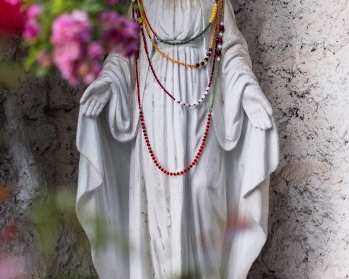 White stone Virgin Mary statue decorated with colorful beads surrounded by pink flowers.