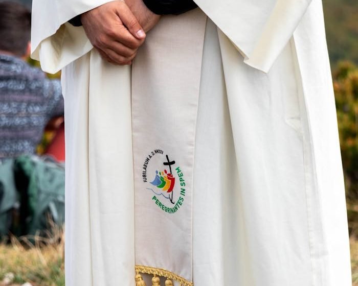 A cleric in a white robe stands outdoors, featuring an embroidered religious symbol.