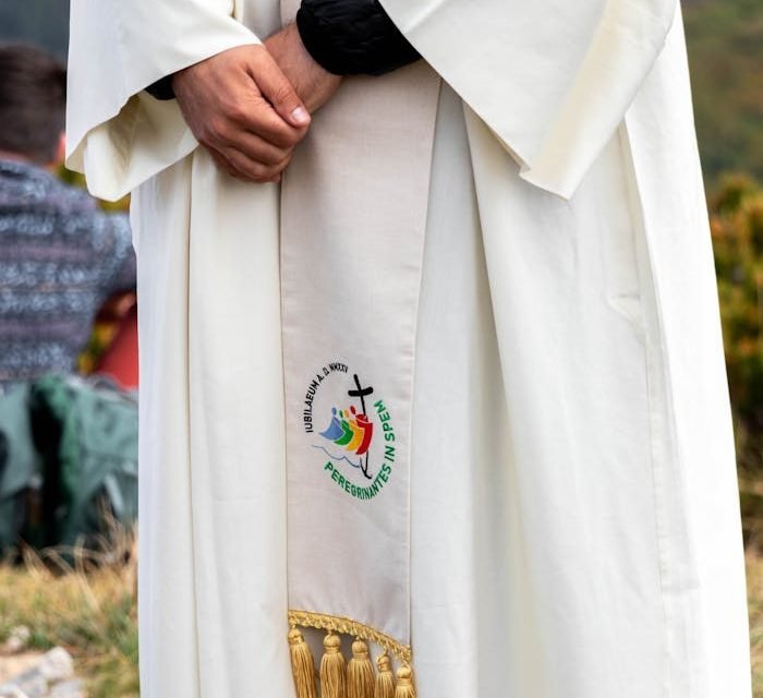 A cleric in a white robe stands outdoors, featuring an embroidered religious symbol.